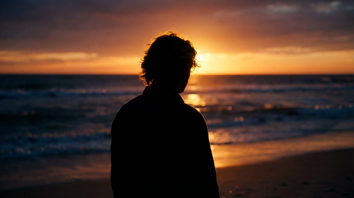 夕暮れの海辺に佇む人物の映画的ショット。逆光によるシルエットと、髪の毛の輪郭を縁取るリムライト。背景には沈みゆく太陽と波の反射。被写界深度（ボケ量）を活かしたシネマティックな表現、実写的なリアルさ。